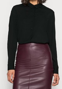 Schwarzes Langarmshirt mit Knopfverschluss, kombiniert mit einem taillierten bordeauxfarbenen Lederrock, der eine glatte Textur und subtile Nahtdetails aufweist.