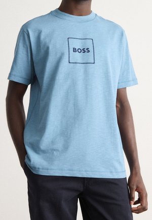 Man wearing light blue short-sleeve T-shirt with dark blue square and "BOSS" text centered, paired with dark pants against plain background.