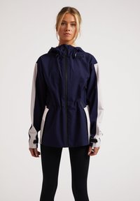 Chaqueta impermeable azul marino y blanca con capucha, cintura elástica y bolsillos laterales. Incluye cremalleras y puños ajustables para mayor comodidad.