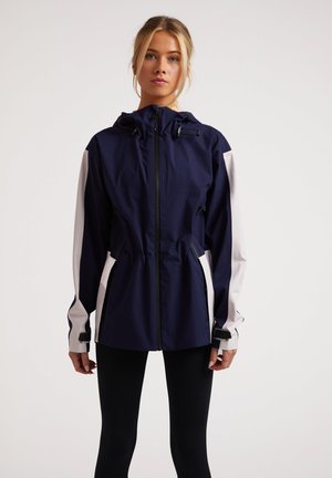 Giacca impermeabile blu navy e bianca con cappuccio, vita elastica e tasche laterali. Presenta cerniere e polsini regolabili per il massimo comfort.