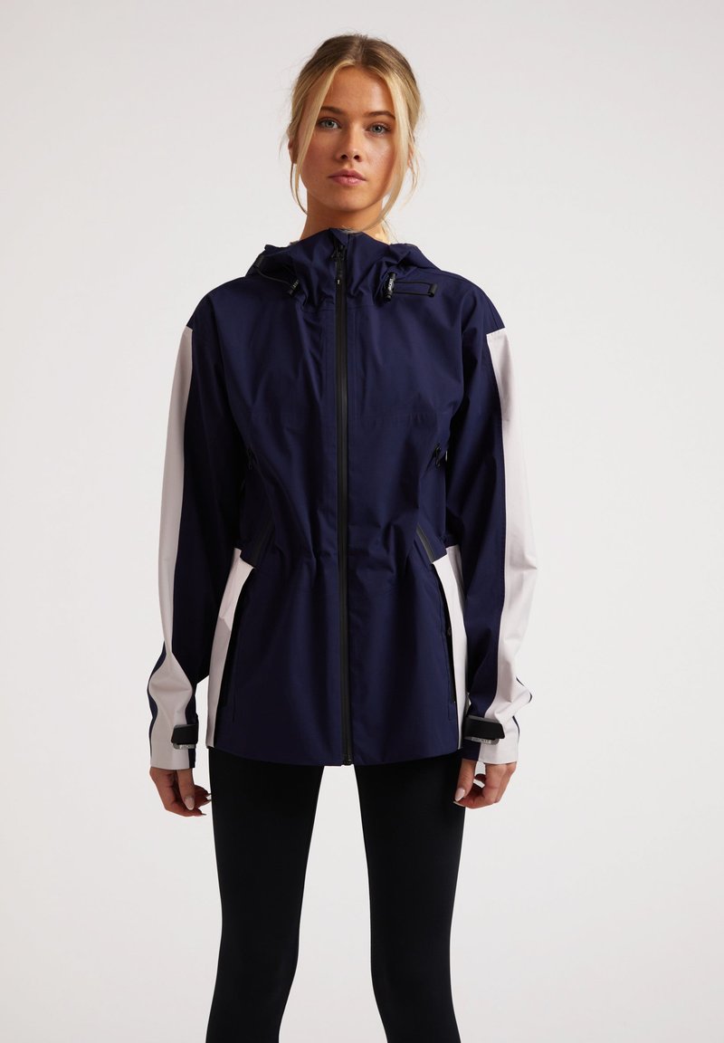 Chaqueta impermeable azul marino y blanca con capucha, cintura elástica y bolsillos laterales. Incluye cremalleras y puños ajustables para mayor comodidad.