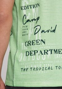 Zelený pruhovaný tielko s čiernym textom a grafikou, vrátane fráz ako "Camp David" a "Zelené oddelenie." Hladká textúra.