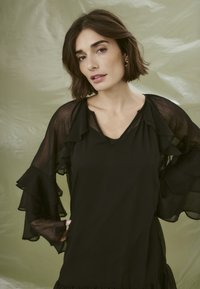 Svart blus med genomskinliga volangärmar. V-halsdesign, mjuk textur och lager av detaljer vid ärmsluten. Enkel och elegant silhuett.