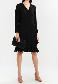 Sort slå-om-kjole med lange ærmer og flæset kant. Akkompagneret af en sort clutch og nude høje hæle. Glat tekstur.