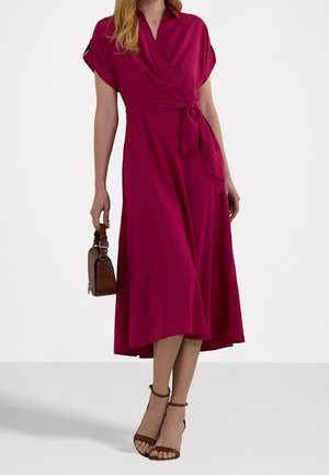 Mujer con un vestido cruzado color rosa intenso hasta la rodilla, de mangas cortas, sosteniendo un pequeño bolso marrón y calzando tacones marrones con correa en el tobillo.