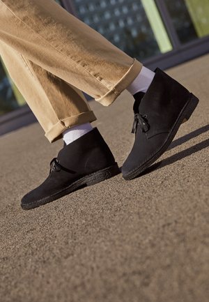 Botas de ante negras hasta el tobillo con una suela texturizada, presentan una punta redondeada y un cierre de cordones, combinadas con pantalones caqui enrollados y calcetines blancos.