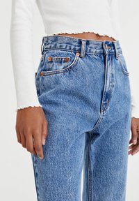 Korkeavyötäröiset siniset farkut, joissa on kulunut viimeistely, viiden taskun malli, kupariset yksityiskohdat ja kontrastiväriset tikkaukset. Valkoinen ribattu pitkähihainen toppi.