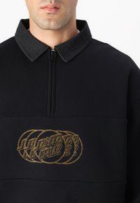 Suéter negro con un cuello con cremallera, que presenta un cuello de tela gris oscuro y detalles del logo bordado en dorado en el pecho.