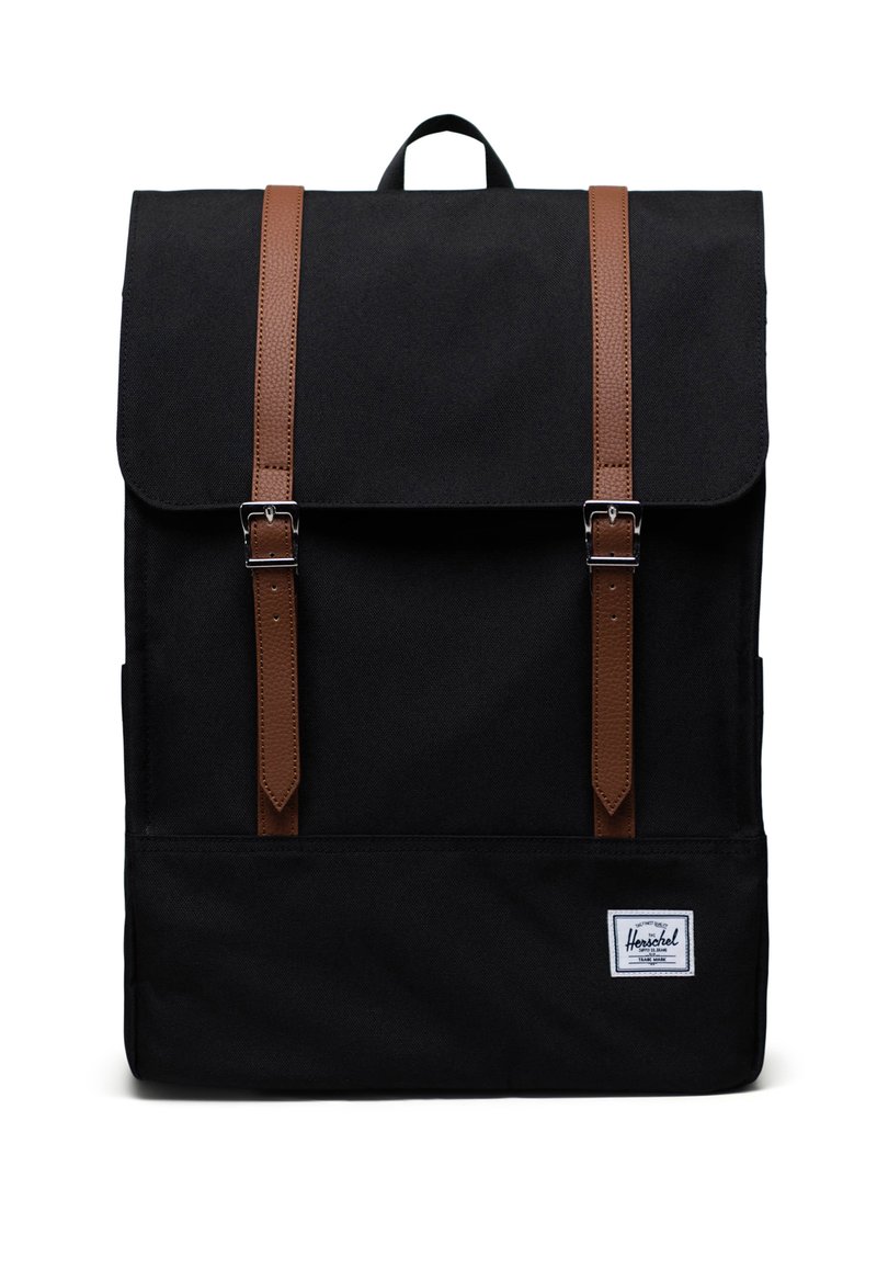 Sac à dos en toile noire avec des sangles en cuir marron, des boucles métalliques et une étiquette logo blanche à l'avant. Il dispose d'une poignée supérieure pour le portage.