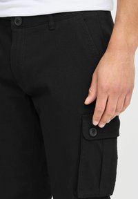 Pantalons cargo noirs avec fermeture à bouton, dotés d'une poche latérale avec un rabat et un détail de bouton, et fabriqués dans un tissu lisse et texturé.