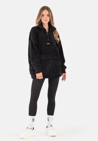 Czarny polarowy pulower z ćwierć zamkiem, z haftem "COZY" na piersi, noszony na czarnych legginsach i białych sneakersach.