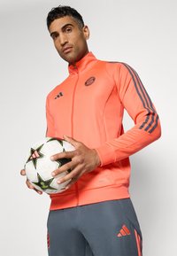 Adidas orangenes Trainingsjackett mit schwarzen Akzenten, hohem Kragen und Logo. Das Model hält einen gemusterten weißen und schwarzen Fußbal und trägt graue Hosen.