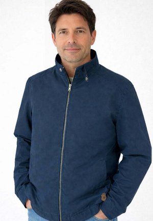 Homme portant une veste bleu marine zippée avec un col montant, mains dans les poches, sur un fond blanc uni.