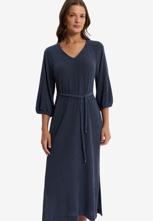 Kobieta ubrana w granatową, prążkowaną sukienkę midi z długim rękawem, dekoltem w serek i wiązanym paskiem w talii na tle jednolitej, białej ściany.