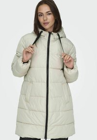 Manteau en crème matelassé avec capuche, fermeture éclair noire, cordon de serrage et deux poches latérales ; présente des panneaux horizontaux cousus et des poignets élastiques.