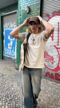 Gaiši beidži t-krekls, vaļīgas izbalējušas džinsi un tumša cepure ar Coca-Cola logo; roku turēts šalle; pilsētas vide ar flīzētām sienām.