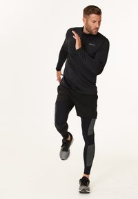 Sort langærmet træningsbluse med halvlukning, parret med sorte shorts og teksturerede marinefarvede leggings, der har grå detaljer og sportssko.