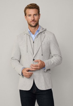 Hellgrauer Sweatshirt-Blazer mit Kapuze, ausgestattet mit einem Reißverschluss, zwei Knöpfen und einer Fronttasche, über einem blauen Hemd getragen.