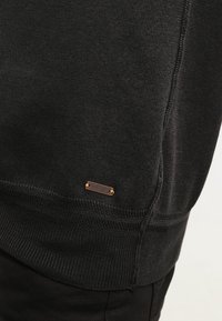 Czarny sweter z dzianiny o ryżowym wzorze i małej miedzianej tabliczce z logo blisko dołu. Posiada luźny krój i minimalistyczny design.