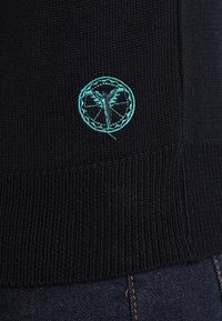 Schwarzer Strickpullover mit strukturiertem Finish und einem türkisfarbigen bestickten Logo einer Pflanze in einem Kreis an der unteren Seite.