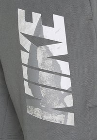 Graue sportliche Jogginghosen mit einem strukturierten Stoff und einem großen, reflektierenden weißen Grafikdesign auf der linken Seite.