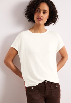 Kvinde med krøllet mørkt hår iført en ensfarvet hvid kortærmet skjorte og mørkebrune bukser, stående med hænderne på hofterne mod en hvid væg.