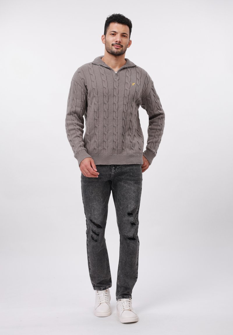 Mężczyzna stoi w szarym swetrze z splotem warkoczowym z zamkiem, czarnych dżinsach z przetarciami i białych sneakersach na jednolitym białym tle.