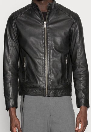 Veste en cuir noire à fermeture éclair avec détails matelassés aux épaules portée avec un pantalon gris, montrant une vue de face du cou aux hanches.