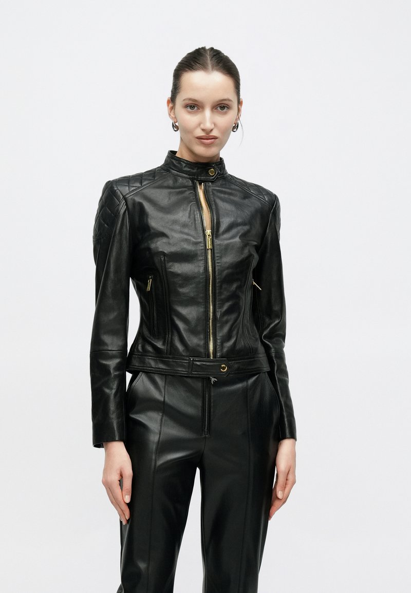 Μαύρο δερμάτινο μπουφάν με ραμμένη καπιτονέ λεπτομέρεια στους ώμους, χρυσά φερμουάρ, ψηλό γιακά και εφαρμοστή σχεδίαση. Διαθέτει ζώνη στη μέση και δύο μπροστινές τσέπες.