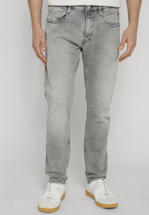 Mann trägt hellgraue, ausgewaschene Jeans und weiße Sneaker mit Gummisohlen, steht vor einem schlichten Hintergrund.