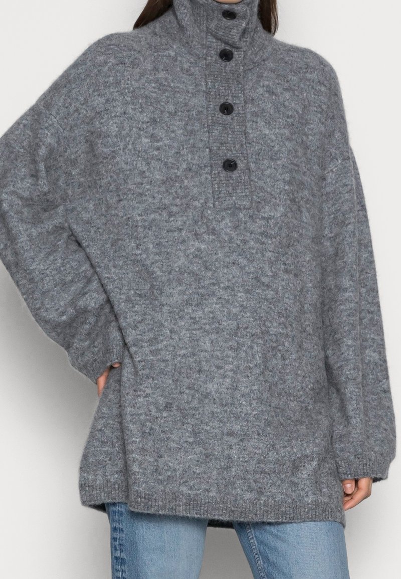 Femme portant un pull oversize en maille grise à col montant et quatre boutons noirs, associé à un jean bleu clair.