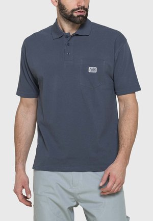 Uomo con barba che indossa una polo blu scuro a maniche corte con tasca sul petto e pantaloni grigio chiaro su sfondo uniforme.