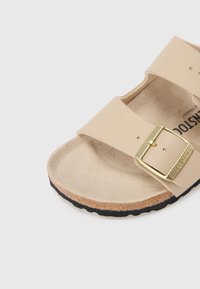 Ανοιχτό μπεζ σανδάλι με ρυθμιζόμενο λουράκι, λεπτομέρεια αγκράφας σε χρυσή απόχρωση, υφασμένο πέλμα και μαύρη λαστιχένια σόλα.