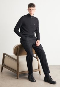 Svart zip-up-genser laget av strikket stoff, kombinert med svarte cargo-bukser med elastisk midje og svarte snørebåndssko. Enkel treskammel med polstret sete.