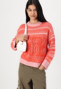 Knitted sweater in coral and pink with textured patterns, ribbed cuffs and neckline. Paired with green cargo pants and a white handbag.