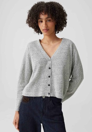 Donna con capelli ricci che indossa un cardigan grigio chiaro abbottonato e jeans scuri a vita alta, in piedi su uno sfondo semplice.