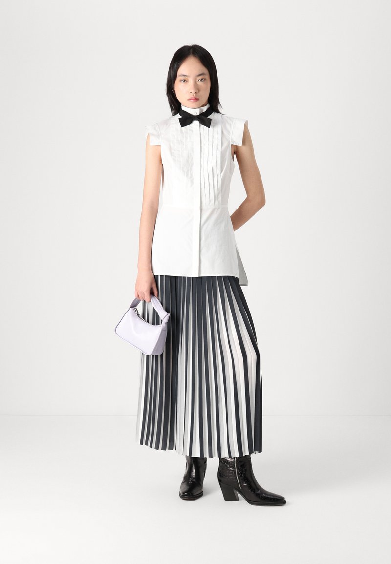 Camisa blanca sin mangas con pliegues, corbata negra de lazo, larga falda plisada gris y blanca, pequeño bolso morado y botas negras de tacón.