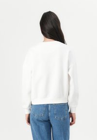 Weißer Sweatshirt mit rundem Halsausschnitt, lässigen Schultern und gerippten Bündchen. Kombiniert mit hochgeschnittenen blauen Jeans.