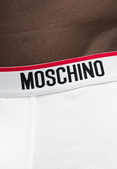 Κοντινό πλάνο στη μέση με σκουρόχρωμο δέρμα που φοράει λευκά εσώρουχα με λευκή ζώνη μέσης που διαθέτει μαύρο κείμενο "MOSCHINO" και κόκκινη λωρίδα στην κορυφή.