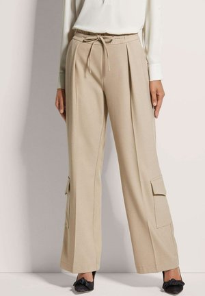 Beige wijde broek gemaakt van zachte stof, met een trekkoord in de taille en zijzakken. Gepaard met zwarte schoenen.