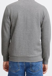 Pull en maille grise avec des manches longues, des poignets ajustés et un ourlet côtelé. Porté avec un jean en denim bleu. Présente un motif côtelé horizontal.