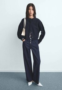 Marineblå stickad kofta med knappar, randig topp, högmidjade mörk denimbyxor och vita spetsiga ballerinas. Modellen håller en liten vit handväska.