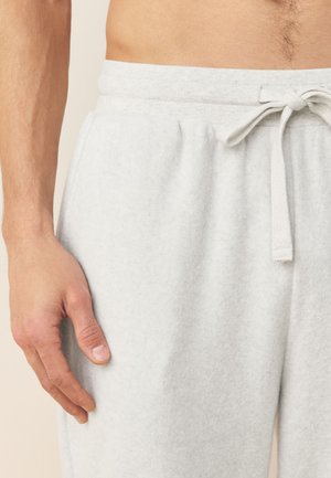 Meessugu torso ja vasak käsi, kandes heledat halli dressipükse, millel on seotud nöör, neutraalsel taustal.