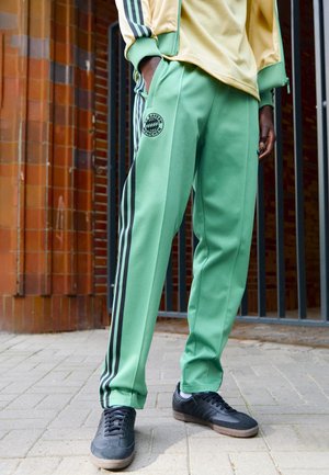 Person iført grønne træningsbukser med sorte striber og Bayern Munchen-logo, kombineret med sorte sneakers og en gul jakke, står på fortov.