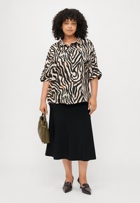 Chemise à manches courtes et à boutons avec un imprimé zèbre noir et beige, associée à une longue jupe noire et un sac vert, avec des chaussures plates noires.