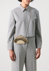 Gold-colored fan-shaped handbag with a patterned frame, held by a person wearing a checked shirt and matching trousers. Chain strap visible.