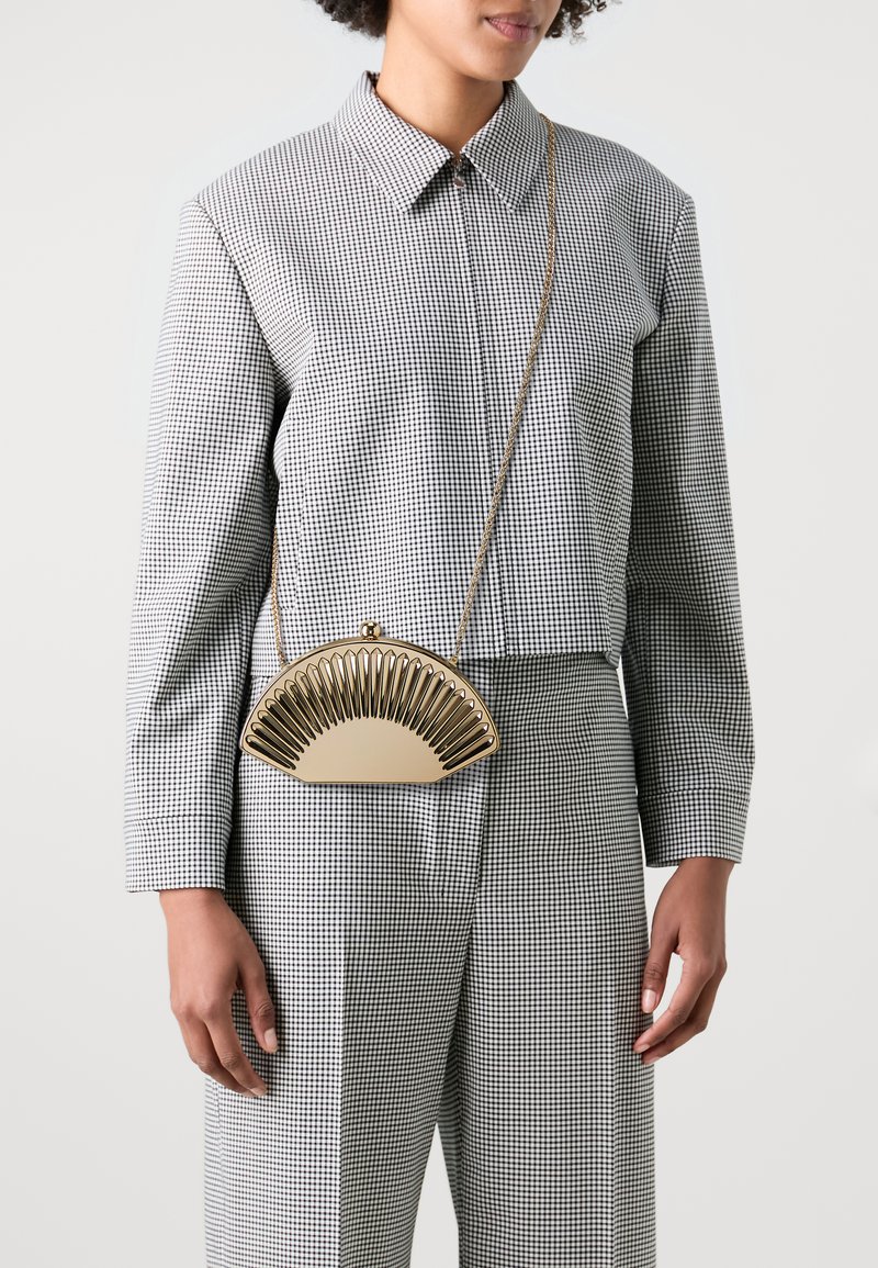 Gold-colored fan-shaped handbag with a patterned frame, held by a person wearing a checked shirt and matching trousers. Chain strap visible.