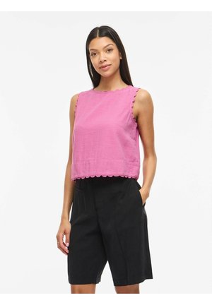 Femme portant un chemisier rose sans manches avec des bords festonnés et un short noir au genou, debout avec une main dans la poche.