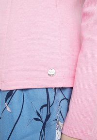 Růžový texturovaný top s dlouhými rukávy a malou kulatou stříbrnou visačkou blízko lemu, doplněný modrými kalhotami s květinovým vzorem.