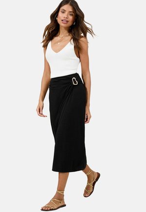 Jupe noire enroulée avec un design tordu, ornée d'une boucle métallique, associée à un débardeur à côtes blanc et des sandales beiges à lanières.